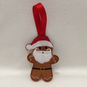 Puede incluir: Adorno navide&ntilde;o de hombre de jengibre de fieltro marr&oacute;n con gorro de Pap&aacute; Noel, barba blanca y cintur&oacute;n negro. El adorno tiene una cinta roja para colgar. El hombre de jengibre tiene costuras blancas y una cara sonriente.