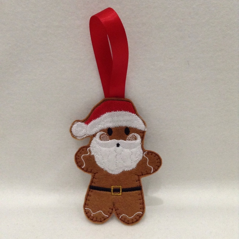 Puede incluir: Adorno navide&ntilde;o de hombre de jengibre de fieltro marr&oacute;n con gorro de Pap&aacute; Noel, barba blanca y cintur&oacute;n negro. El adorno tiene una cinta roja para colgar. El hombre de jengibre tiene costuras blancas y una cara sonriente.