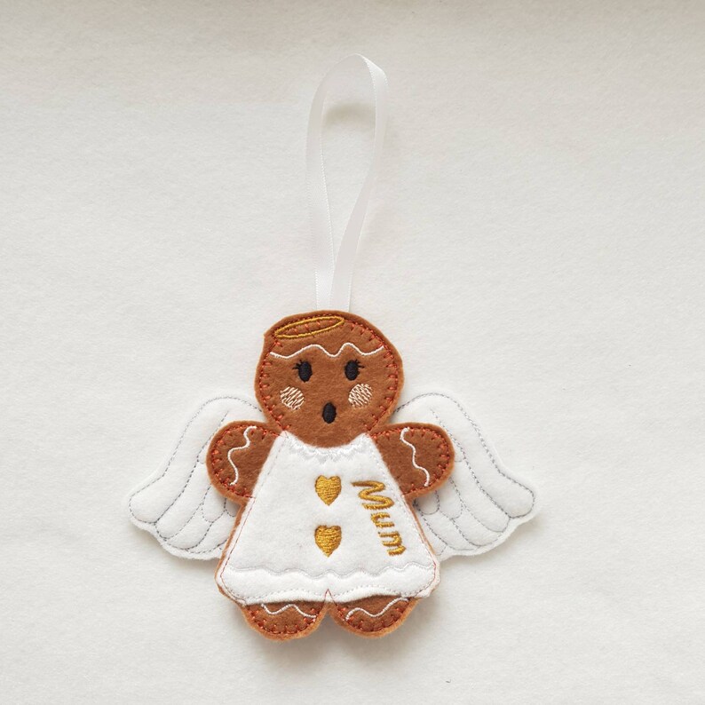 Puede incluir: Adorno de &aacute;ngel de jengibre de fieltro con vestido y alas blancas. La figura de jengibre marr&oacute;n tiene un halo y la palabra "Mum" bordada en dorado. Se adjunta una cinta blanca para colgar.