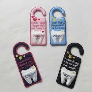 May include: Four tooth fairy door hangers with different colors and names. The hangers are pink, blue, navy blue, and black. Each hanger has a tooth shape with a name on it and the text "Tooth Fairy, Please visit me tonight."
