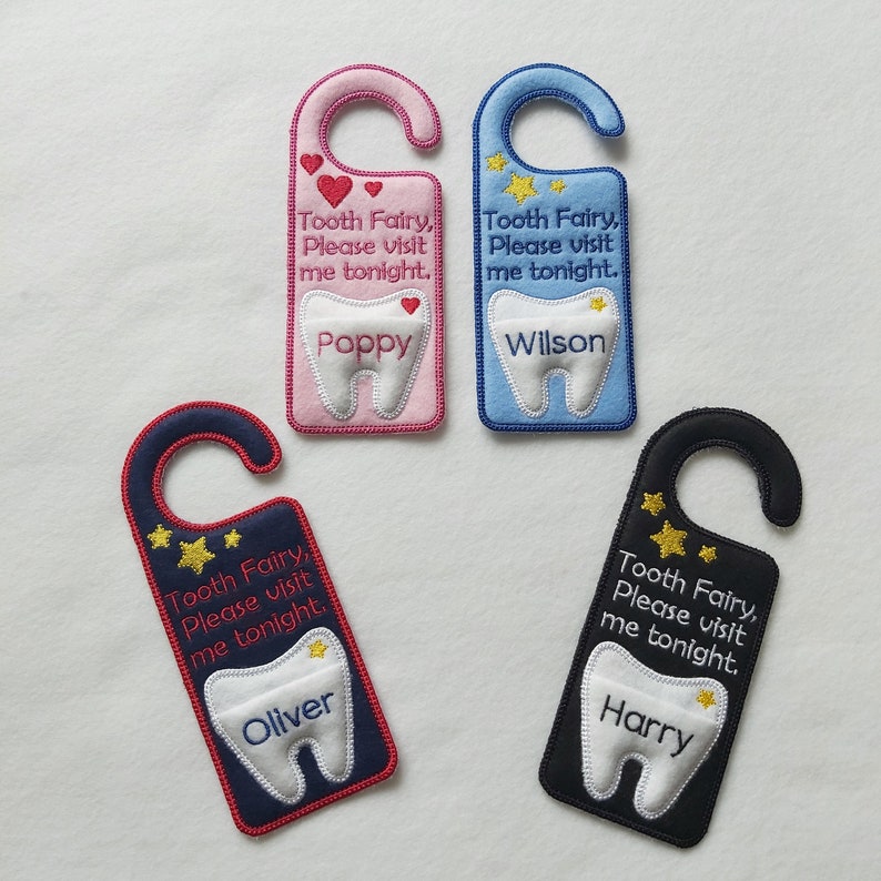 May include: Four tooth fairy door hangers with different colors and names. The hangers are pink, blue, navy blue, and black. Each hanger has a tooth shape with a name on it and the text "Tooth Fairy, Please visit me tonight."