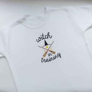 May include: White t-shirt with the embroidered text "witch in training". The design includes a black witch hat, crossed broomsticks, a yellow moon, and two crescent moons. The shirt is made of a soft, white fabric.