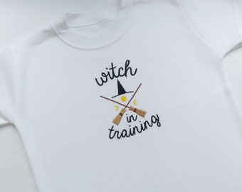 Puede incluir: Camiseta blanca con la frase bordada "witch in training". El diseño incluye un sombrero de bruja negro, escobas cruzadas, una luna amarilla y dos medias lunas. La camiseta está hecha de tela blanca suave.