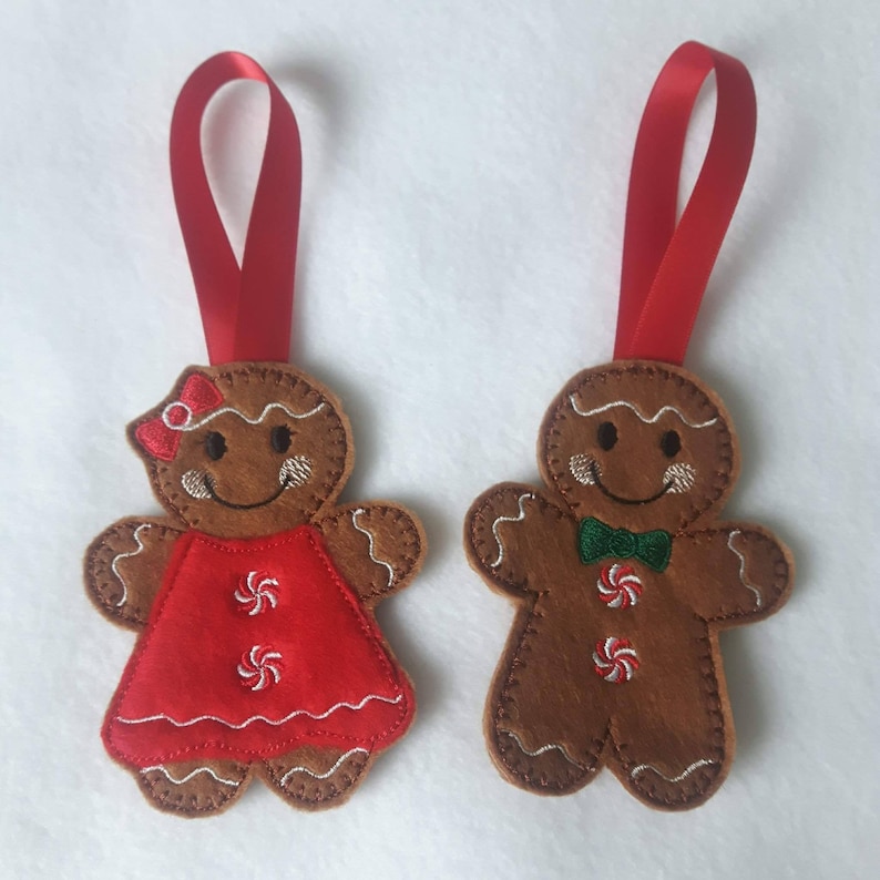 May include: Two gingerbread man ornaments with red ribbon hangers. One wears a red dress with white embroidered details, the other a green bow tie. Both have white icing details and embroidered smiles.