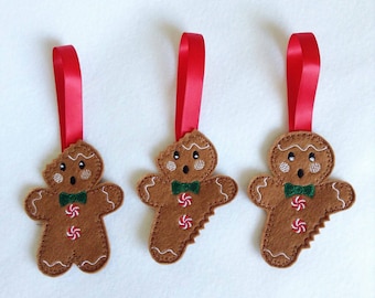 May include: Three brown felt gingerbread man ornaments with red ribbon hangers. Each ornament has a green bow tie, white icing details, and red and white candy accents. The ornaments are on a white background.