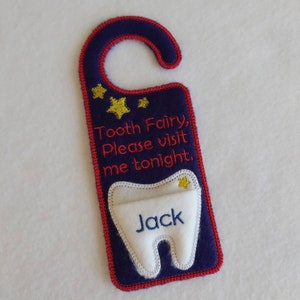 May include: A blue and red door hanger with a white tooth pocket. The tooth pocket has a gold star and the name "Jack" embroidered on it. The door hanger has the text "Tooth Fairy, Please visit me tonight."