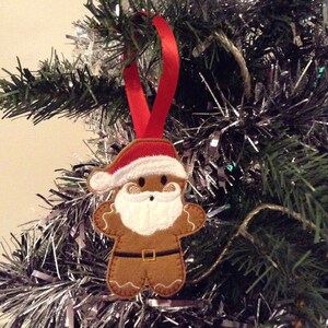 Puede incluir: Adorno navide&ntilde;o de hombre de jengibre con gorro de Pap&aacute; Noel rojo y barba blanca, colgado de una cinta roja. El adorno es marr&oacute;n con detalles blancos y negros, y una hebilla dorada. Cuelga de un &aacute;rbol de Navidad.