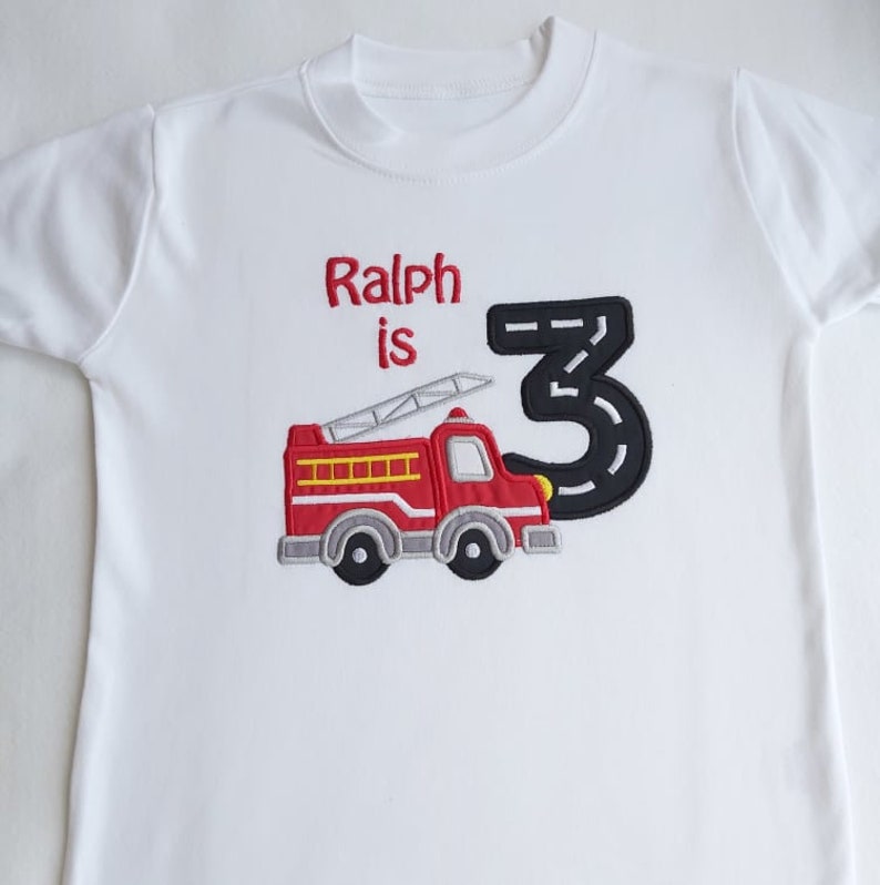 May include: White t-shirt with red and black embroidered fire truck and the text "Ralph is 3" on it.
