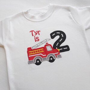 May include: White t-shirt with red, yellow, and black embroidered fire truck and the text "Tyr is 2" in black.