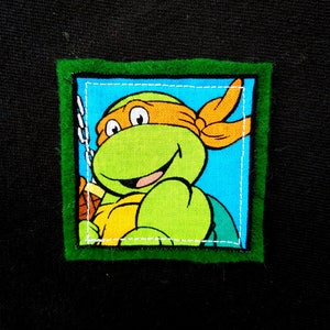 May include: A green, orange, and blue fabric patch with a cartoon image of a Teenage Mutant Ninja Turtle. The patch is surrounded by a green felt border.