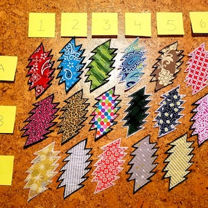 Op de afbeelding: Een verzameling van 18 kleurrijke stoffen patches in de vorm van een bliksem. De patches zijn gerangschikt in rijen en kolommen, waarbij elke rij is gelabeld met een letter (A, B, C) en elke kolom met een nummer (1-6).