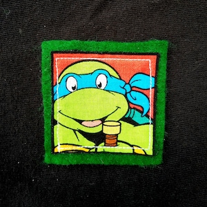 May include: Green felt patch with a red, white, and blue cartoon image of a Teenage Mutant Ninja Turtle wearing a blue bandana and holding a brown stick.