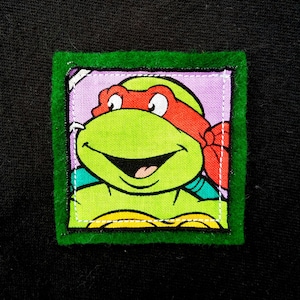 May include: Green felt framed patch with a cartoon image of a Teenage Mutant Ninja Turtle with a red bandana and a green shell. The turtle is smiling with its tongue sticking out.