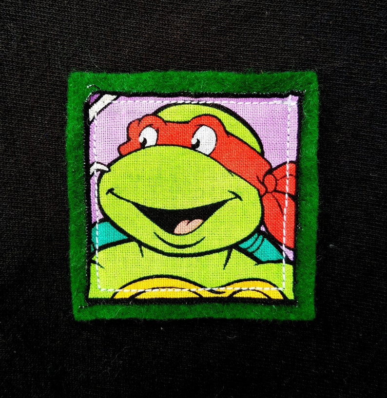 May include: Green felt framed patch with a cartoon image of a Teenage Mutant Ninja Turtle with a red bandana and a green shell. The turtle is smiling with its tongue sticking out.
