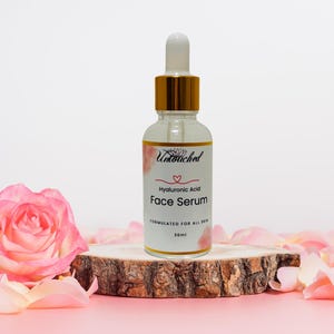 May include: A clear glass bottle of Hyaluronic Acid Face Serum with a gold dropper, resting on a wooden slice. The bottle is labeled with the brand name and product details. Pink rose petals and a pink rose surround the serum.