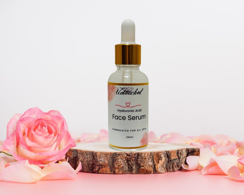 May include: A clear glass bottle of Hyaluronic Acid Face Serum with a gold dropper, resting on a wooden slice. The bottle is labeled with the brand name and product details. Pink rose petals and a pink rose surround the serum.