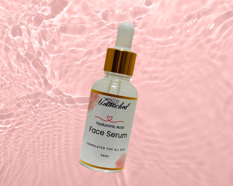 May include: A clear glass bottle of hyaluronic acid face serum with a white and gold cap and dropper. The label reads "Untouched" and "Face Serum". The background is a pink, rippled surface.