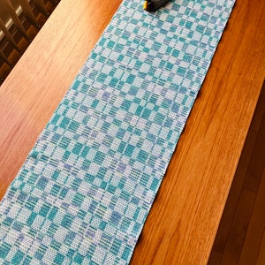 Peut inclure: Un chemin de table à carreaux blancs et turquoise avec une figurine de canard décorative sur le dessus. Le chemin de table est sur une table en bois.