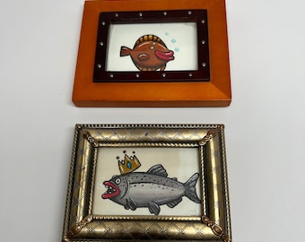 May include: Two framed illustrations of fish. The top frame is brown with a gold border and features a brown fish with a red mouth. The bottom frame is silver with a gold border and features a gray fish with a gold crown and red mouth.