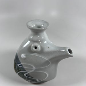 May include: A ceramic sake bottle with a unique design. The bottle is primarily gray with white, blue, and black accents. It features a spout and a small opening at the top. The bottle has a whimsical, artistic appearance.