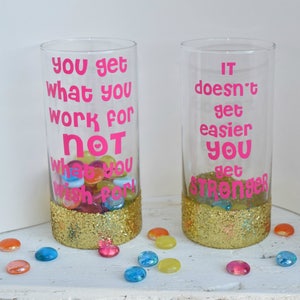 Puede incluir: Dos tarros de vidrio transparente con purpurina dorada alrededor de la parte superior. Los tarros tienen citas motivacionales en letras rosas. El tarro de la izquierda dice "You get what you work for not what you wish for!" El tarro de la derecha dice "It doesn't get easier you get stronger."
