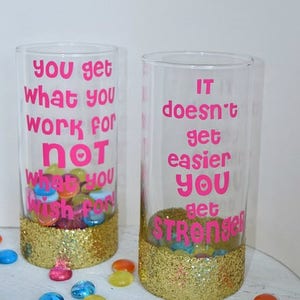 Puede incluir: Dos vasos de vidrio transparente con purpurina dorada en el fondo. Los vasos tienen letras de vinilo rosa con citas motivacionales. El primer vaso dice "You get what you work for not what you wish for". El segundo vaso dice "It doesn't get easier you get stronger".