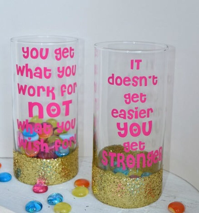 Puede incluir: Dos vasos de vidrio transparente con purpurina dorada en el fondo. Los vasos tienen letras de vinilo rosa con citas motivacionales. El primer vaso dice "You get what you work for not what you wish for". El segundo vaso dice "It doesn't get easier you get stronger".