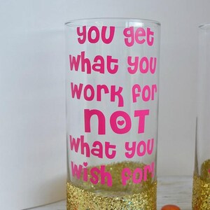 Puede incluir: Un vaso transparente con un borde de purpurina dorada y el texto "you get what you work for not what you wish for" en letras rosas.