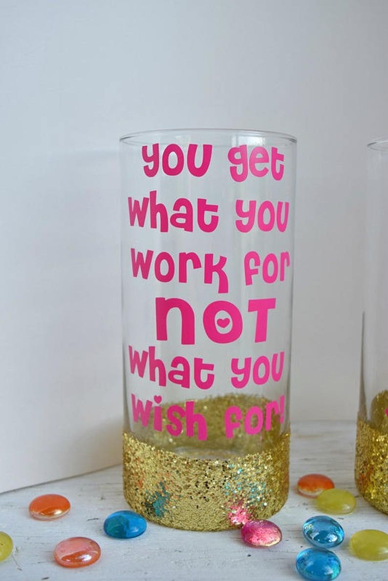 Puede incluir: Un vaso transparente con un borde de purpurina dorada y el texto "you get what you work for not what you wish for" en letras rosas.