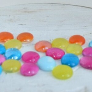 Puede incluir: Surtido de canicas de vidrio de colores en tonos de azul, verde, amarillo, naranja, rosa y rojo. Las canicas est&aacute;n esparcidas sobre una superficie blanca.