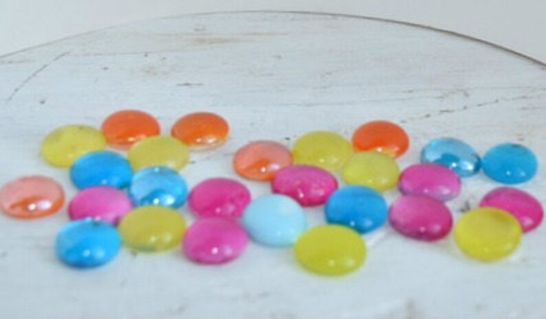 Puede incluir: Surtido de canicas de vidrio de colores en tonos de azul, verde, amarillo, naranja, rosa y rojo. Las canicas est&aacute;n esparcidas sobre una superficie blanca.