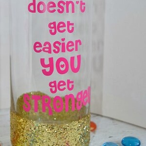 Puede incluir: Un vaso de vidrio transparente con un borde de purpurina dorada y el texto "IT doesn't get easier YOU get STRONGER" en letras rosas.