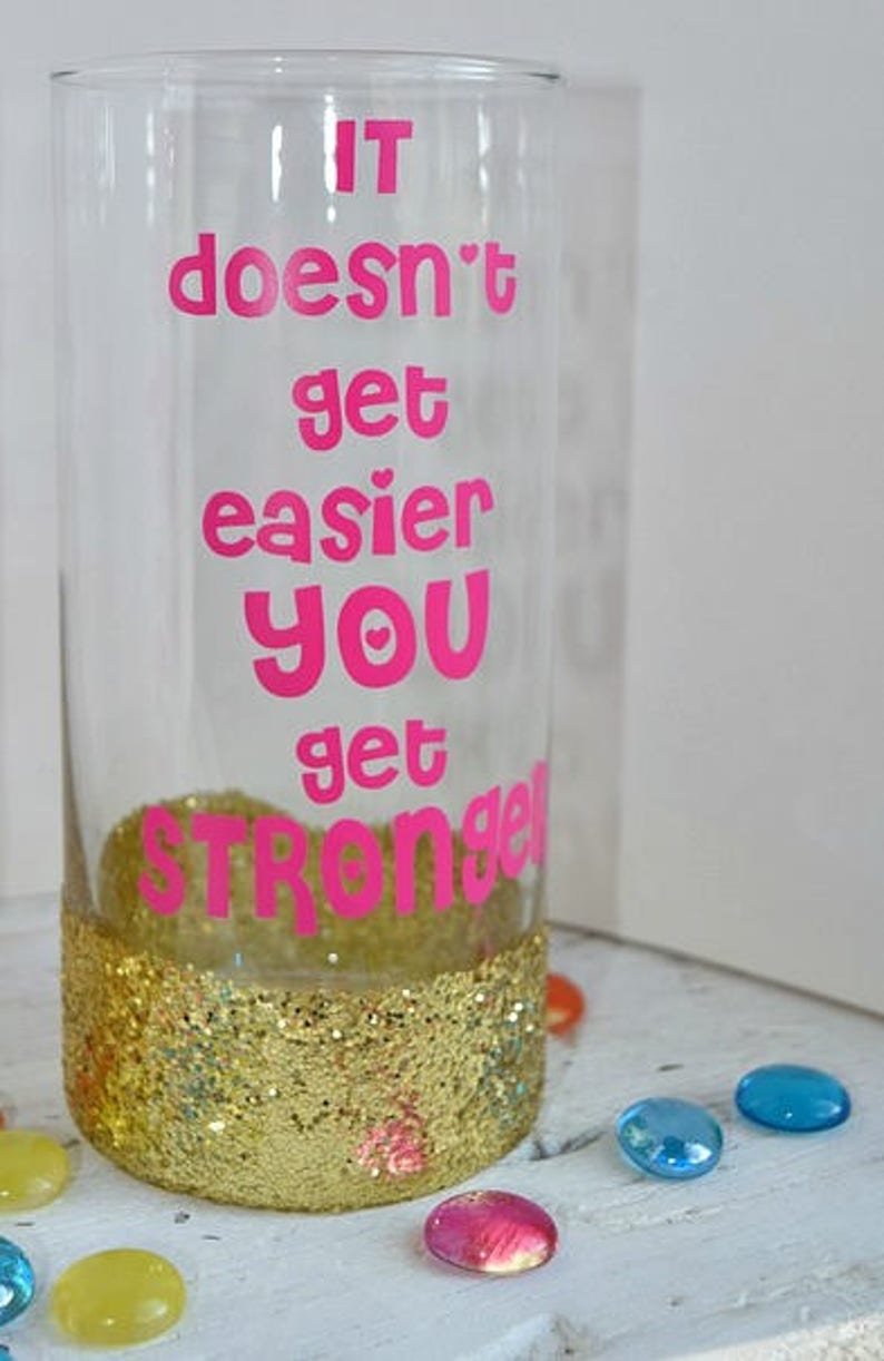 Puede incluir: Un vaso de vidrio transparente con un borde de purpurina dorada y el texto "IT doesn't get easier YOU get STRONGER" en letras rosas.