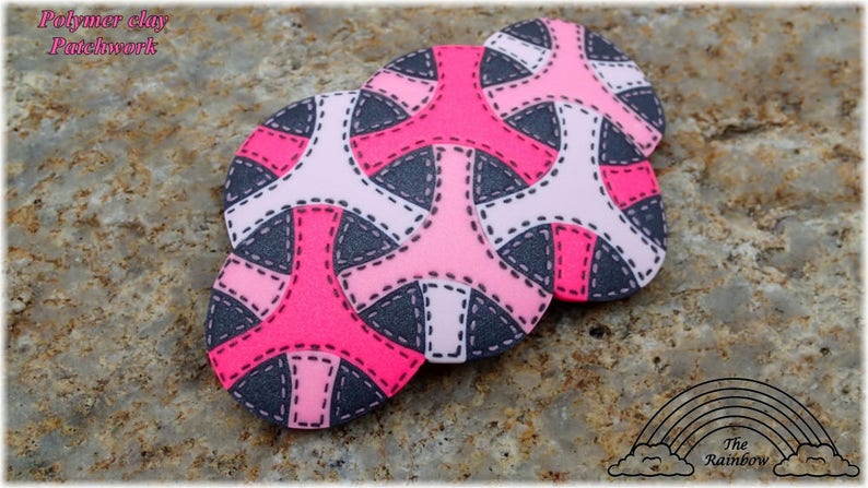 May include: Three pink, white, and gray polymer clay circles with a geometric pattern. The circles are arranged in a stack, with the largest circle on the bottom and the smallest circle on top. The circles have a stitched design around the edges.
