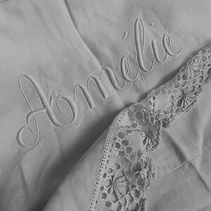 May include: A white linen blanket with the name "Amélie" embroidered in cursive script. The blanket has a delicate lace trim along one edge.