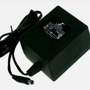 Op de afbeelding: Een zwarte rechthoekige AC-stroomadapter met een bevestigd netsnoer en een cilindrische stekker. Een zwart label op de bovenzijde bevat in het wit gedrukte technische specificaties, inclusief de UL- en CSA-certificeringssymbolen.
