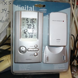 May include: A silver digital thermometer with a wireless remote sensor. The thermometer displays the temperature in Fahrenheit and the time. The remote sensor has a red light on it.