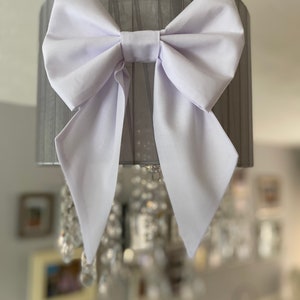 May include: A white fabric bow tied around a grey lampshade. The lampshade is made of a sheer fabric with a pleated design.