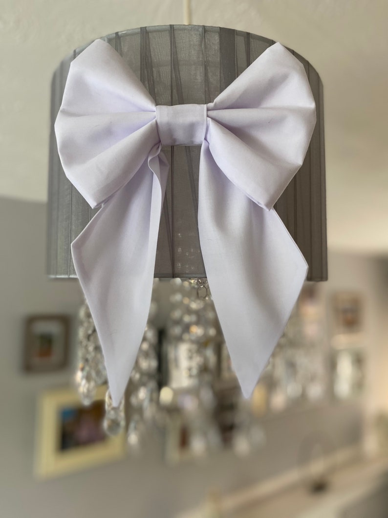 May include: A white fabric bow tied around a grey lampshade. The lampshade is made of a sheer fabric with a pleated design.