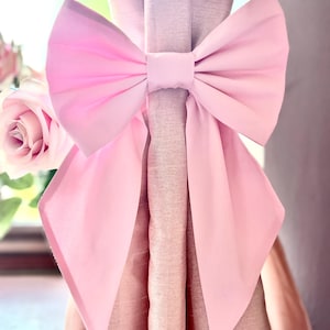 May include: A large, pale pink bow tied around a curtain. The bow is made of a soft, flowing fabric and is centered on the curtain. A pink rose is visible in the background, adding a touch of elegance.