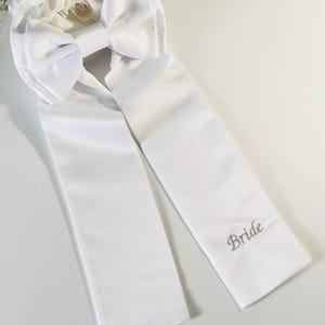 Könnte beinhalten: Weißes Satin-Brautschärpe mit großer Schleife und langen Bändern. Auf einem Band ist das Wort "Bride" in silberner Schrift gestickt. Perfekt für eine Hochzeit.