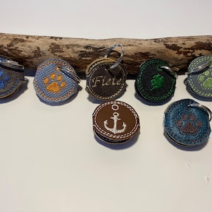 Peut inclure: Six porte-clés différents avec des motifs variés. Les porte-clés sont en cuir brun avec des accents de tissu de différentes couleurs. Les motifs comprennent une empreinte de patte, une ancre, un trèfle et le mot "Fiete".