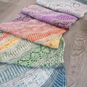 Puede incluir: Una bufanda de punto degradada en arcoíris con un patrón de encaje. La bufanda está hecha de hilo suave y tiene una sensación delicada y aireada.