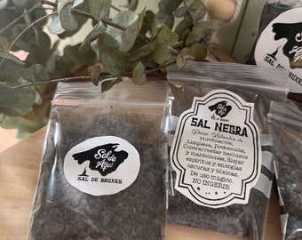 May include: Two small bags of black salt, one labeled "Sal de Bruxes" and the other "Sal Negra". Both bags are on a wooden surface with green eucalyptus leaves in the background.