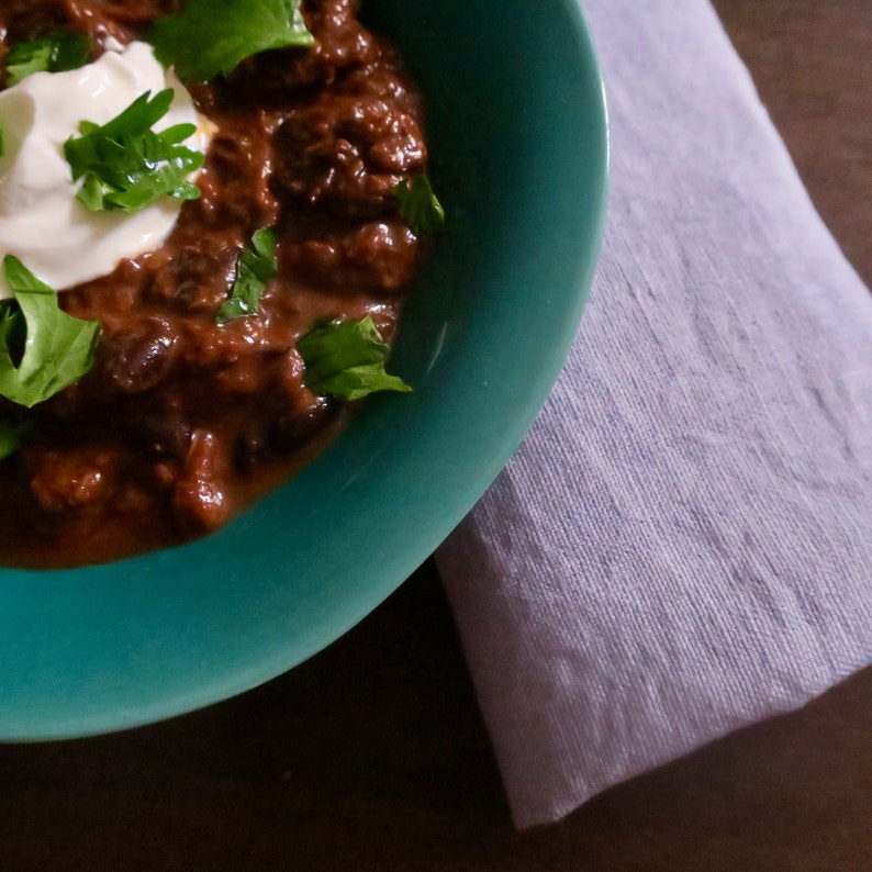 May include: A close-up of a bowl of chili with a dollop of sour cream and chopped cilantro on top. The bowl is teal blue and the chili is a dark brown color.