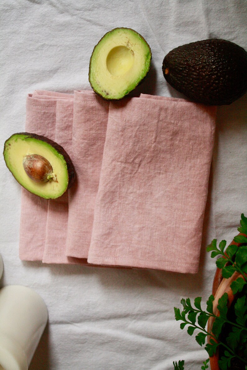 May include: A set of four pink linen napkins folded on a white tablecloth with two halved avocados on the table.