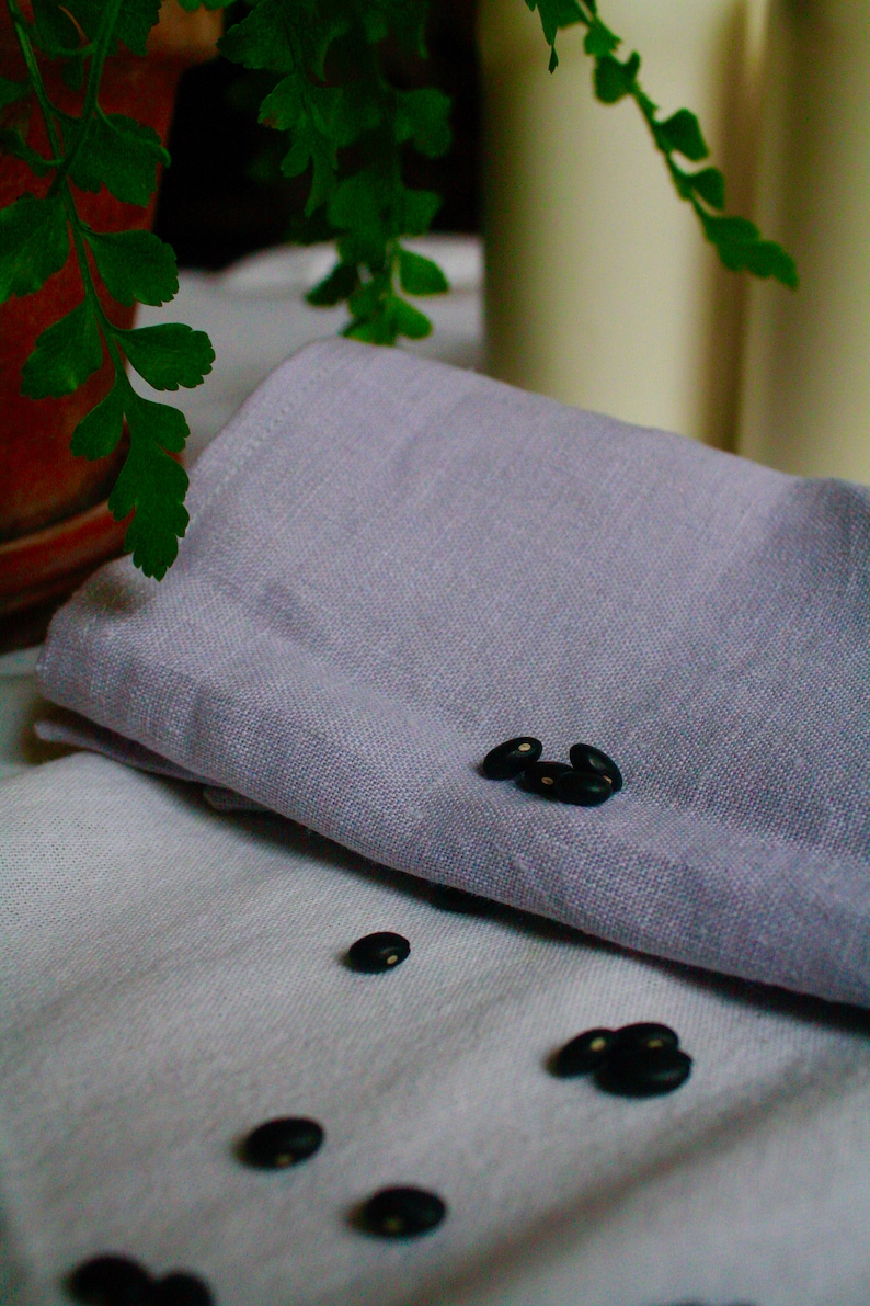 May include: Black beans scattered on a white linen tablecloth with a folded purple linen napkin on top.