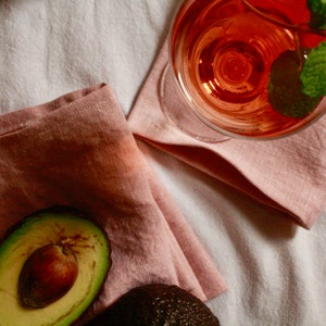 May include: A close-up of a half avocado with a pit on a pink linen napkin. A glass of pink wine with a sprig of mint is in the background.