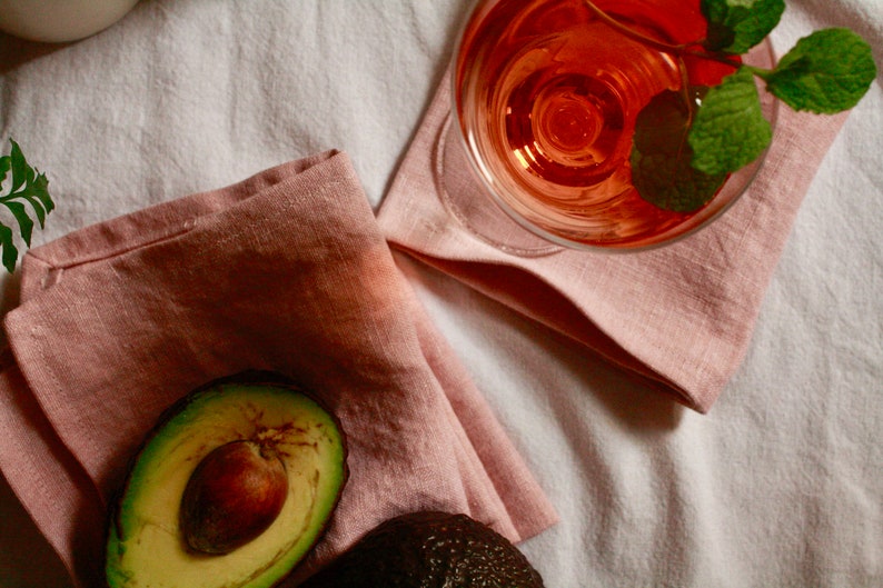 May include: A close-up of a half avocado with a pit on a pink linen napkin. A glass of pink wine with a sprig of mint is in the background.