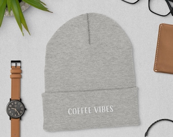May include: A light gray beanie with the embroidered text "COFFEE VIBES" in white. The beanie is folded at the brim. A brown leather watch, black glasses, and a small brown wallet are also visible.